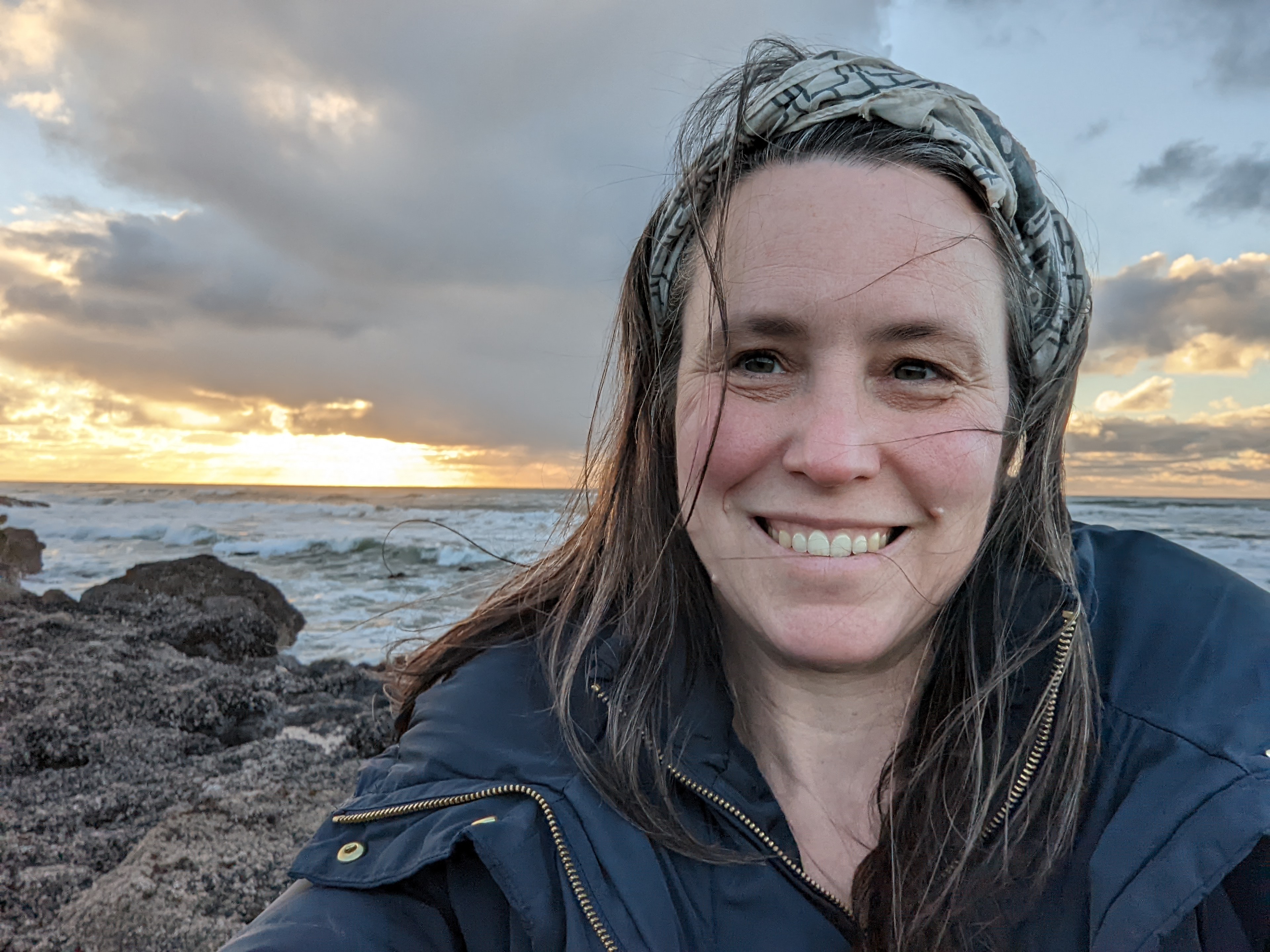 close up photo of a white female smiling. She is wearing a comfy jacket and has straight long brown hair blowing in the wind. Behind her is a backdrop of an ocean with breaking waves.